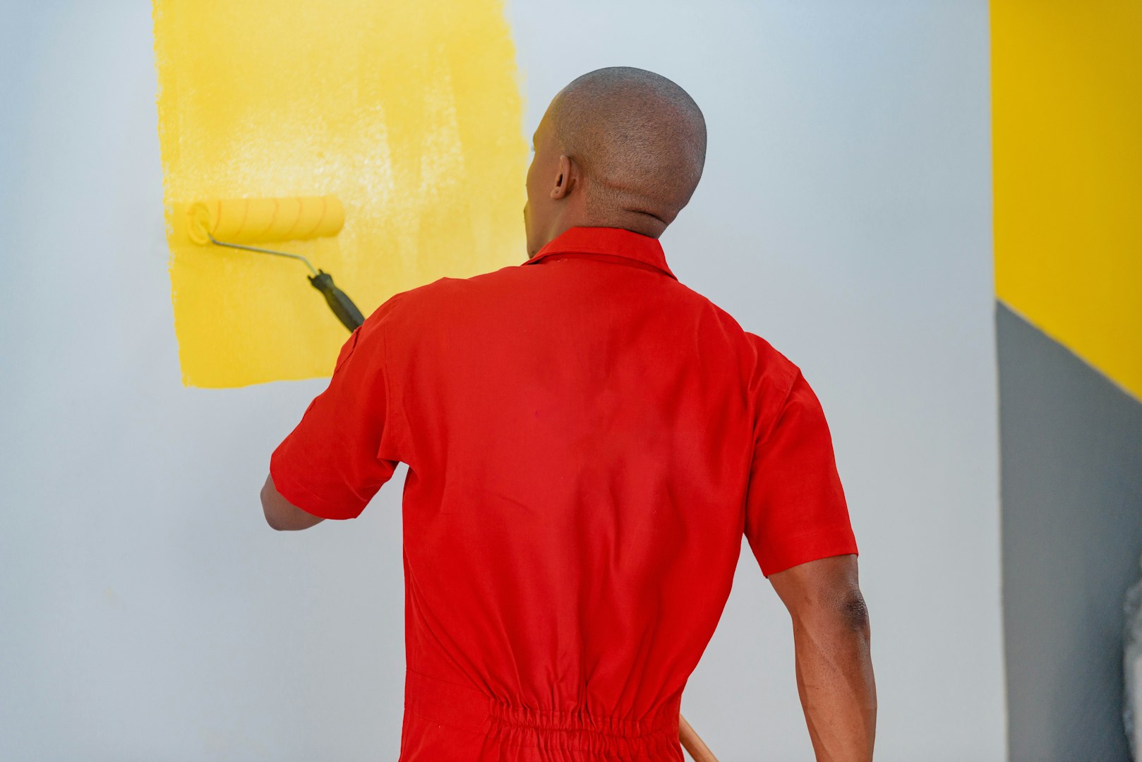 A woman wearing a blue apron and yellow gloves is cleaning a surface in a sunlit room filled with plants and decorative items. She is focused on her task, surrounded by a bright and cheerful environment.